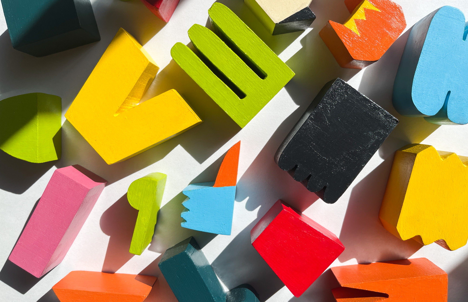 A collection of brightly colored wooden blocks in various geometric shapes, including rectangles, triangles, and abstract forms, scattered across a white surface. The blocks are painted in vivid hues of yellow, green, red, blue, pink, black, and orange, casting sharp shadows due to strong overhead lighting.